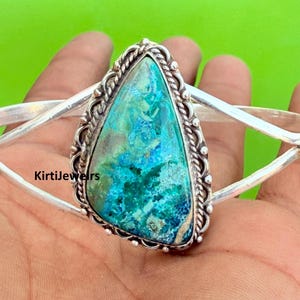 May include: A silver cuff bracelet with a large, triangular teal and blue stone. The stone is set within a decorative, textured border. The bracelet is held in a hand against a green background. The text "Kirtijewels" is visible.