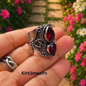 May include: A wide silver ring featuring two vertically stacked, pear-shaped red gemstones set in metal bezels. The band is decorated with intricate silver wire scrollwork, beaded accents, and a twisted rope border. The text KirtiJewelrs is visible.