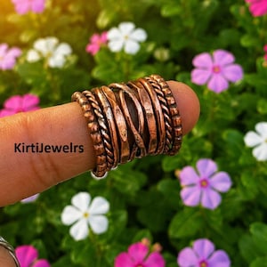 May include: A wide copper ring featuring a multi-band design with various textures, including twisted rope patterns and hammered finishes. The text KirtiJewelrs is visible on the side of the ring, which is displayed on a finger against a floral background.