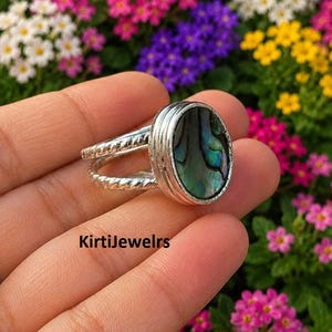 Puede incluir: Un anillo de plata con una incrustación ovalada de concha de abulón en tonos verdes y azules iridiscentes. La banda consta de dos hilos de plata trenzados que se unen en un engaste liso para la piedra. El texto KirtiJewelrs es visible.