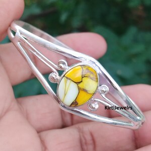 Könnte beinhalten: Ein silbernes Armband mit einem zentralen ovalen gelb-weißen Stein mit dunkler Maserung. Das Metallband hat ein geteiltes Design mit kleinen silbernen Kugeln, die die Steinfassung betonen. Der Text KirtiJewelrs ist auf dem Band zu sehen.