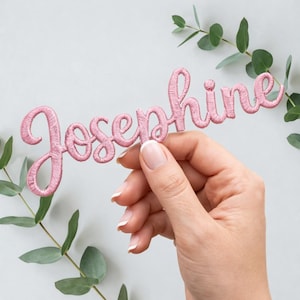 Puede incluir: Un parche bordado rosa con la palabra "Josephine" en cursiva. Lo sostiene una mano con uñas cuidadas, con hojas de eucalipto de fondo. El parche es de un tono rosa claro.