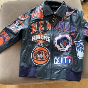 Op de afbeelding: Een donker marineblauw leren jack met meerdere geborduurde patches van New Yorkse sportteams, waaronder de Knicks en Rangers, met de tekst NEW YORK en KITH prominent op de voorzijde en mouwen, en een zwart label aan de kraag bevestigd.