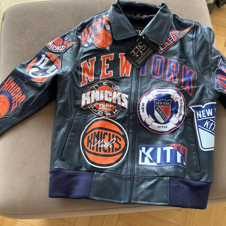 Op de afbeelding: Een donker marineblauw leren jack met meerdere geborduurde patches van New Yorkse sportteams, waaronder de Knicks en Rangers, met de tekst NEW YORK en KITH prominent op de voorzijde en mouwen, en een zwart label aan de kraag bevestigd.