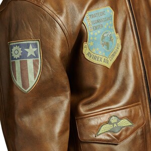 May include: Close-up of a brown leather jacket with multiple patches. The jacket features a patch with the text "STRATEGIC RECONNAISSANCE CENTER" and "AVIREX AIR COMMAND". Another patch displays a star and stripes design. A pocket with a winged emblem is also visible.