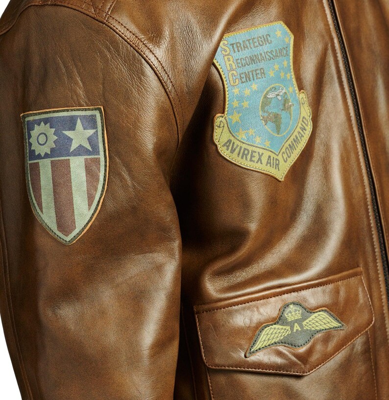May include: Close-up of a brown leather jacket with multiple patches. The jacket features a patch with the text "STRATEGIC RECONNAISSANCE CENTER" and "AVIREX AIR COMMAND". Another patch displays a star and stripes design. A pocket with a winged emblem is also visible.