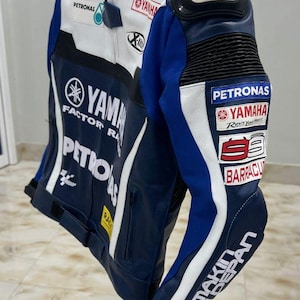 Puede incluir: Una chaqueta de cuero para motocicleta en azul, blanco y negro con logotipos de Yamaha Factory Racing, Petronas y Bridgestone. La chaqueta incluye un protector de hombro plateado, bolsillos con cremallera y varios logotipos de patrocinadores.
