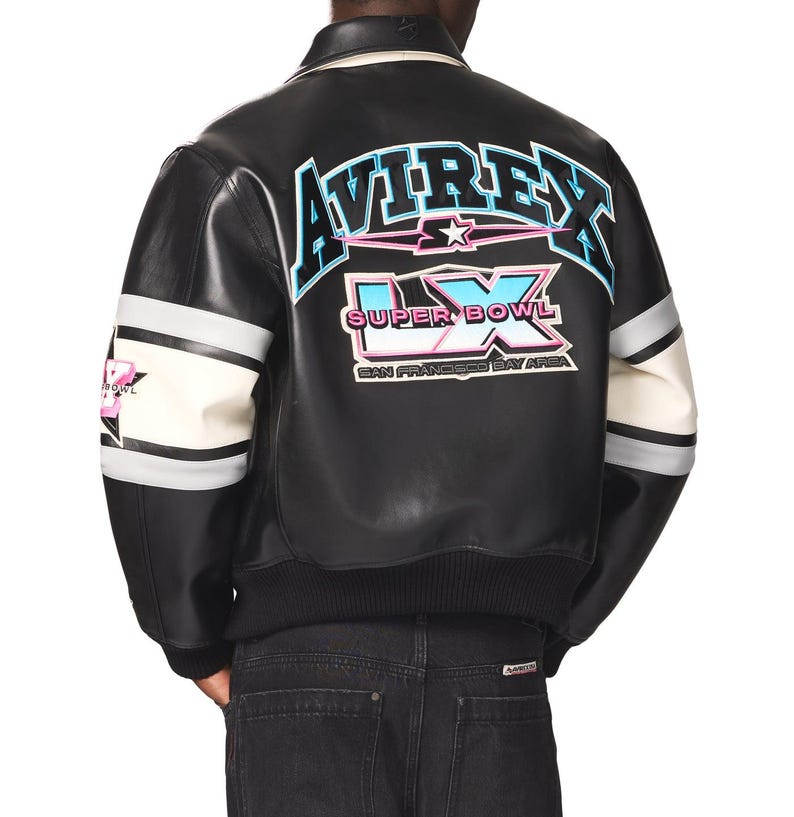 Peut inclure: Veste de bombardier en cuir noir avec des manches ray&eacute;es blanches et grises. Le dos pr&eacute;sente le texte "AVIREX" en bleu et rose, ainsi que "SUPER BOWL LX SAN FRANCISCO BAY AREA". La veste a un ourlet et un col c&ocirc;tel&eacute;s.