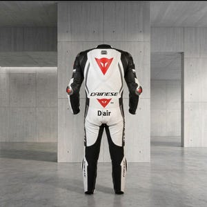 Puede incluir: Traje de carreras de motociclismo blanco y negro con detalles en rojo. El traje presenta el logotipo "DAINESE" y la palabra "Dair". Tiene protecciones en hombros y codos. El traje est&aacute; dise&ntilde;ado para la velocidad y la seguridad.