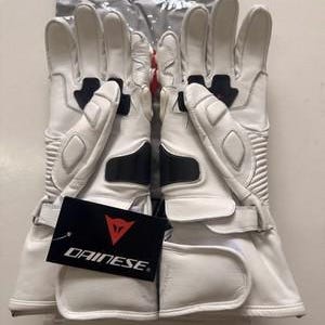 K&ouml;nnte beinhalten: Paar wei&szlig;e Motorradhandschuhe mit schwarzen Akzenten und einem Handgelenkriemen. Die Handschuhe haben ein Etikett mit dem Dainese-Logo. Die Handschuhe sind aus Leder und haben Schutzelemente.