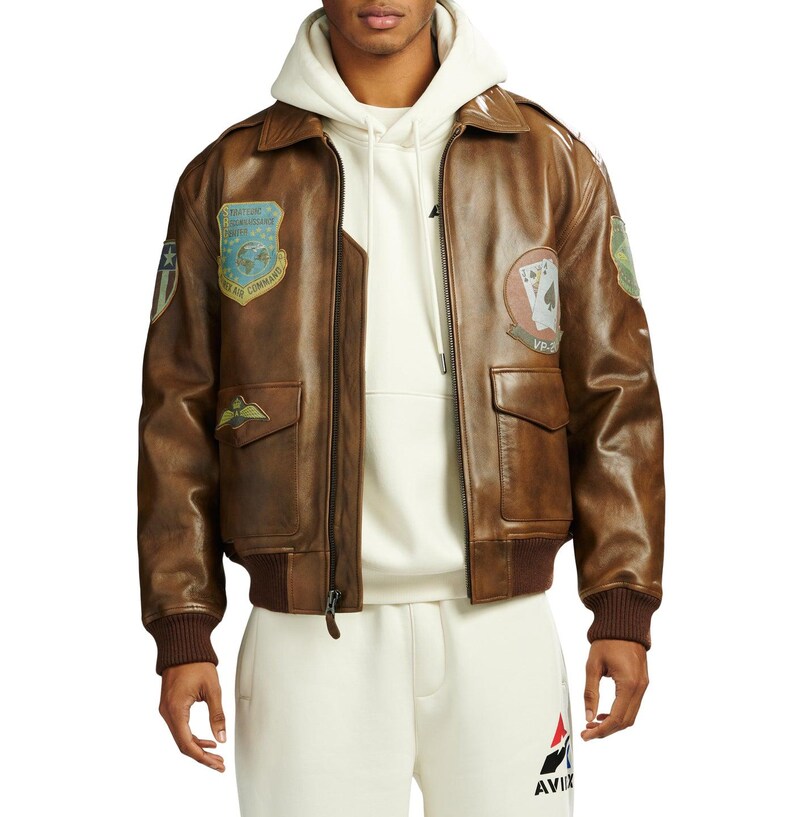 May include: Brown leather bomber jacket with patches. The jacket has a zip closure, two front pockets, and a ribbed collar and cuffs. Patches include military-style emblems and text. The jacket is worn over a white hoodie and sweatpants.