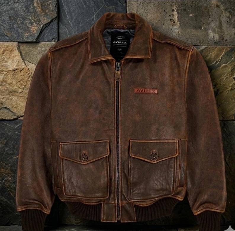 May include: A brown leather jacket with a zip-up front, two front pockets, and a collar. The jacket has a distressed look and the brand name "AVIREX" is visible on a patch. The jacket is displayed against a textured stone background.