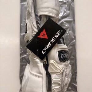 K&ouml;nnte beinhalten: Wei&szlig;e Motorradhandschuhe aus Leder mit Schutzpolstern und einem schwarzen Etikett mit dem roten Dainese-Logo. Die Handschuhe befinden sich in einer transparenten Plastikt&uuml;te.