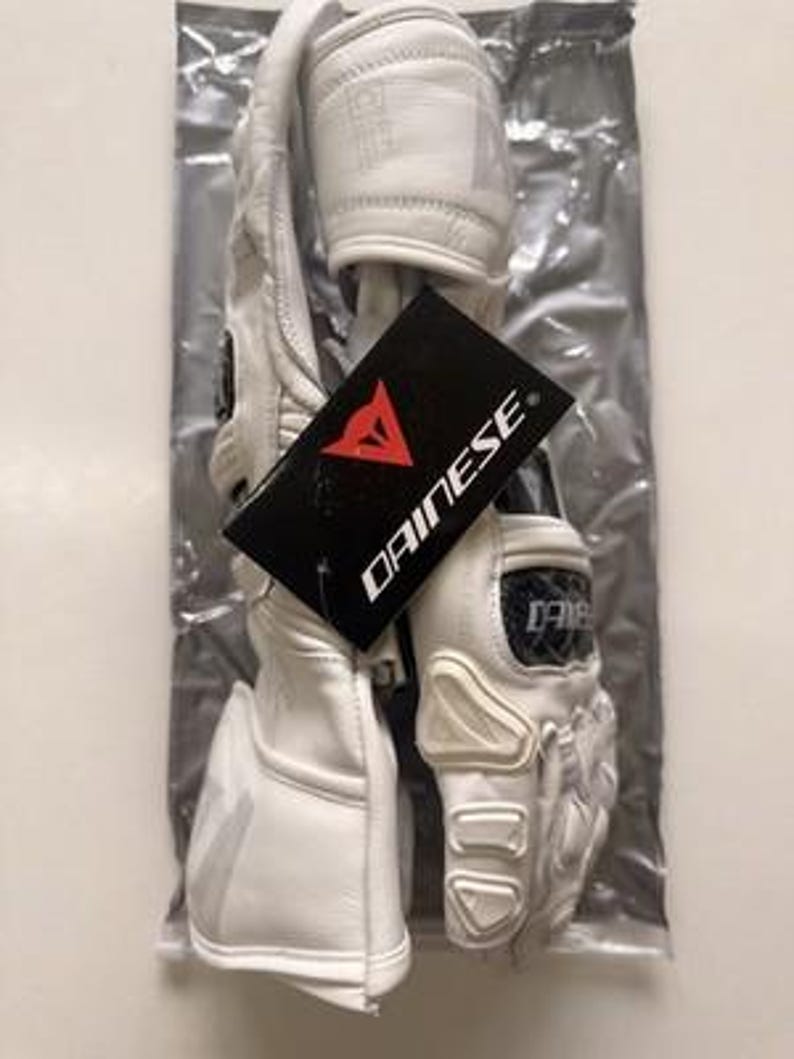 K&ouml;nnte beinhalten: Wei&szlig;e Motorradhandschuhe aus Leder mit Schutzpolstern und einem schwarzen Etikett mit dem roten Dainese-Logo. Die Handschuhe befinden sich in einer transparenten Plastikt&uuml;te.
