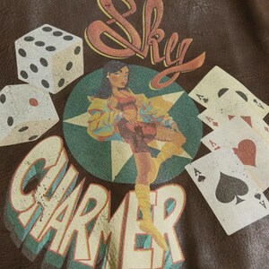 May include: Brown leather-like material with a graphic design featuring dice, playing cards, and a woman. The text "Sky Charmer" is prominently displayed. The artwork has a vintage, retro aesthetic.