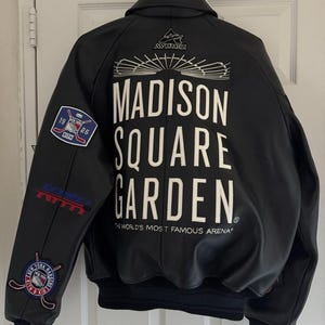 Op de afbeelding: Een zwart leren varsity-jack met witte tekst op de achterkant die luidt MADISON SQUARE GARDEN THE WORLD'S MOST FAMOUS ARENA. Het jack bevat een Avirex-logo, een New York Rangers Kith-patch en aanvullende sportieve branding.
