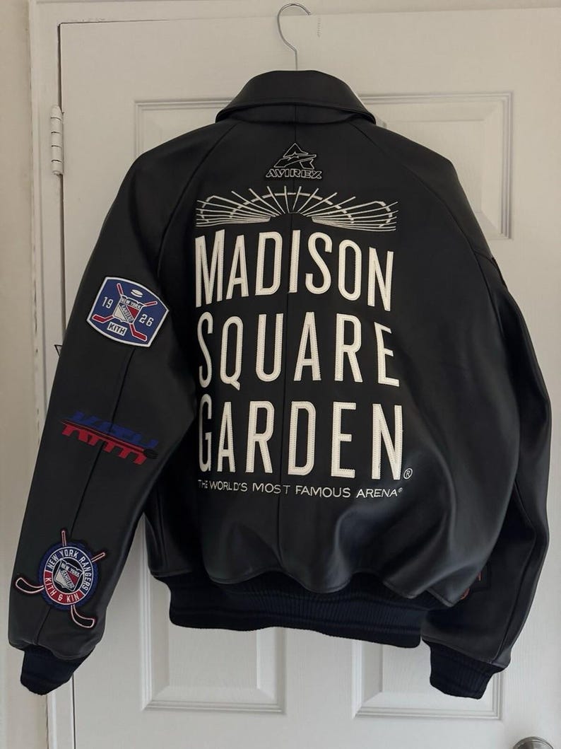 Op de afbeelding: Een zwart leren varsity-jack met witte tekst op de achterkant die luidt MADISON SQUARE GARDEN THE WORLD'S MOST FAMOUS ARENA. Het jack bevat een Avirex-logo, een New York Rangers Kith-patch en aanvullende sportieve branding.