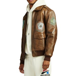May include: Brown leather bomber jacket with patches, including a VP-21 patch with playing cards. The jacket has a ribbed collar and cuffs, and two front pockets. The Avirex logo is visible on the pants.