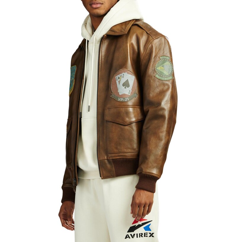 May include: Brown leather bomber jacket with patches, including a VP-21 patch with playing cards. The jacket has a ribbed collar and cuffs, and two front pockets. The Avirex logo is visible on the pants.