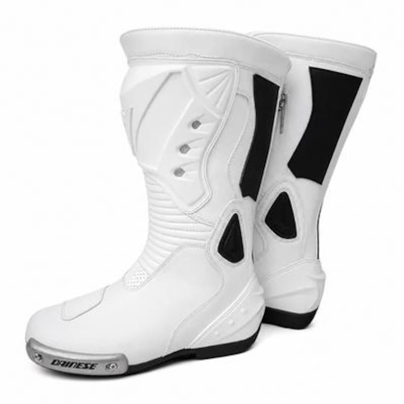 K&ouml;nnte beinhalten: Wei&szlig;e Motorradstiefel mit schwarzen Akzenten. Die Stiefel haben ein stromlinienf&ouml;rmiges Design mit Schutzfunktionen, einschlie&szlig;lich verst&auml;rkter Paneele und einem Rei&szlig;verschluss. Der Markenname "Dainese" ist sichtbar.