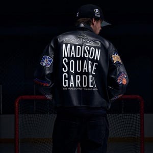 Op de afbeelding: Een zwart leren varsity-jack met witte tekst op de achterkant die luidt MADISON SQUARE GARDEN THE WORLD'S MOST FAMOUS ARENA, voorzien van diverse geborduurde sportteam-patches op de mouwen en een zwarte baseballpet op het hoofd.