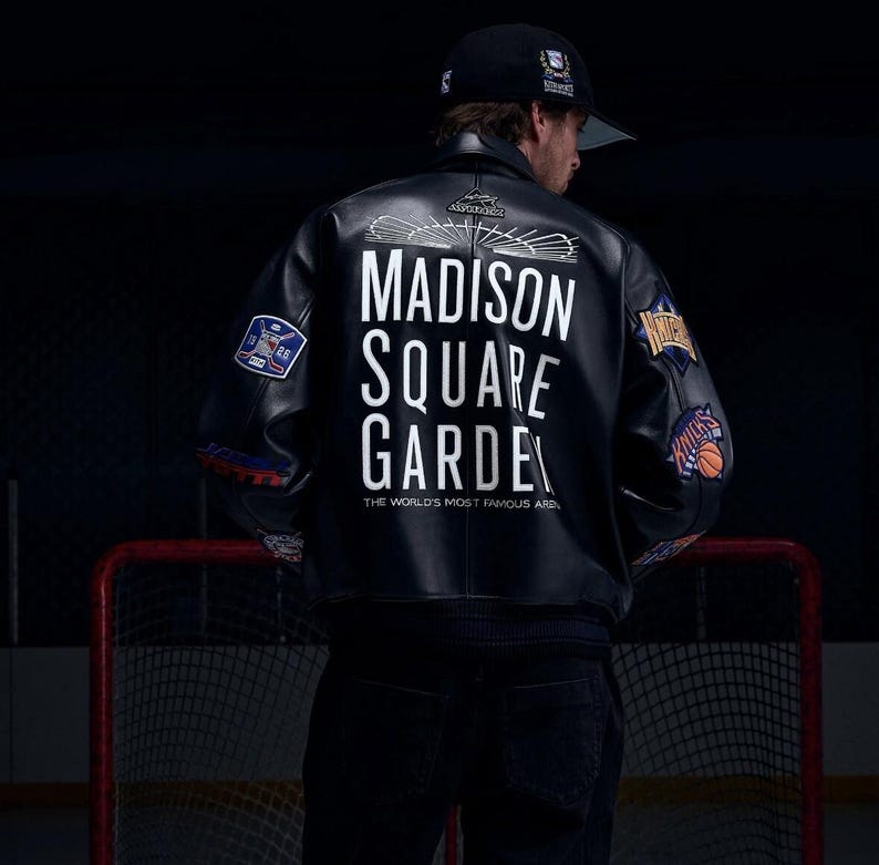 Op de afbeelding: Een zwart leren varsity-jack met witte tekst op de achterkant die luidt MADISON SQUARE GARDEN THE WORLD'S MOST FAMOUS ARENA, voorzien van diverse geborduurde sportteam-patches op de mouwen en een zwarte baseballpet op het hoofd.