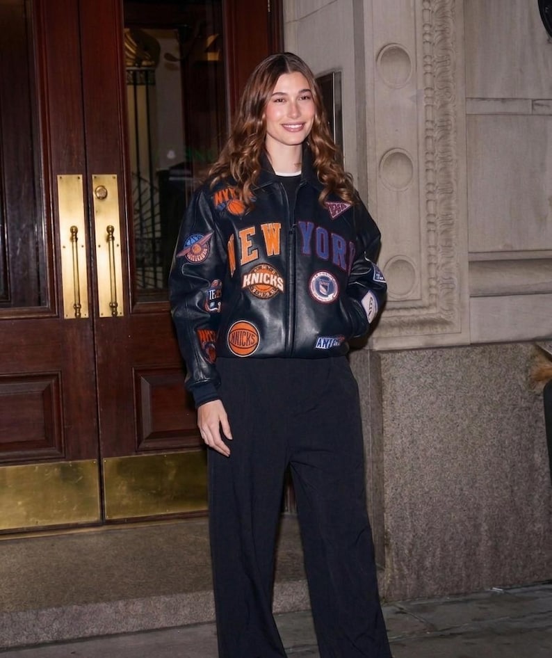 Op de afbeelding: Een donkerblauwe leren bomberjack met diverse geborduurde patches en de tekst NEW YORK in oranje en paarse letters. Het jack heeft een kraag, lange mouwen met geribbelde boorden en een ritssluiting aan de voorzijde, gecombineerd met een zwarte broek.