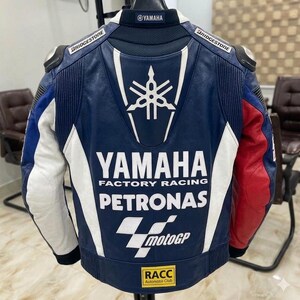 Puede incluir: Una chaqueta de carreras de cuero azul, blanca y roja con el logotipo de Yamaha, el texto Yamaha Factory Racing, la marca Petronas, el logotipo de MotoGP y un parche del RACC Automobil Club en la espalda. Logos de Bridgestone en los hombros.