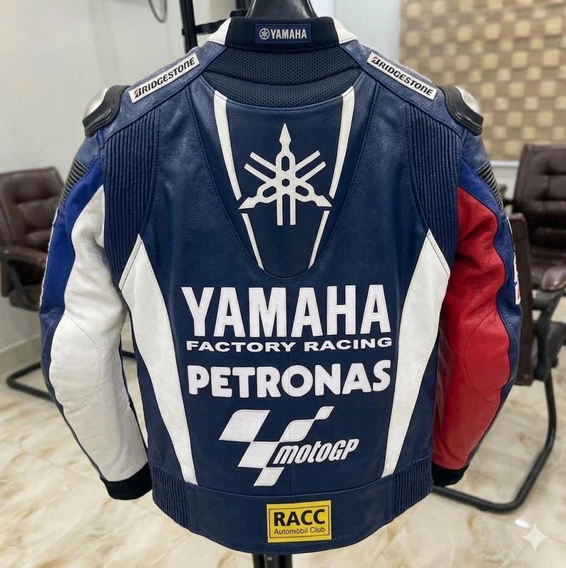 Puede incluir: Una chaqueta de carreras de cuero azul, blanca y roja con el logotipo de Yamaha, el texto Yamaha Factory Racing, la marca Petronas, el logotipo de MotoGP y un parche del RACC Automobil Club en la espalda. Logos de Bridgestone en los hombros.