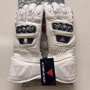 K&ouml;nnte beinhalten: Wei&szlig;e Motorradhandschuhe mit Schutzelementen. Die Handschuhe haben harte Kn&ouml;chelsch&uuml;tzer und verst&auml;rkte Handfl&auml;chen. Der Markenname "DAINESE" ist auf einem schwarzen Etikett an den Handschuhen zu sehen. Die Handschuhe befinden sich in einer transparenten Plastikt&uuml;te.