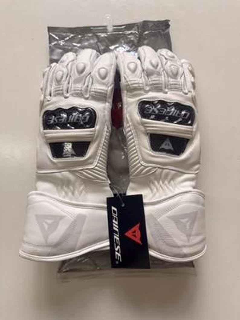 K&ouml;nnte beinhalten: Wei&szlig;e Motorradhandschuhe mit Schutzelementen. Die Handschuhe haben harte Kn&ouml;chelsch&uuml;tzer und verst&auml;rkte Handfl&auml;chen. Der Markenname "DAINESE" ist auf einem schwarzen Etikett an den Handschuhen zu sehen. Die Handschuhe befinden sich in einer transparenten Plastikt&uuml;te.