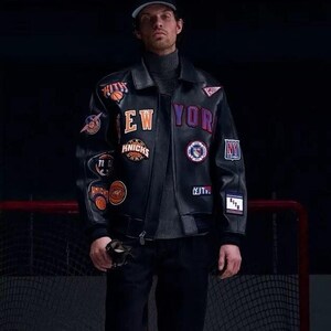 Op de afbeelding: Een zwart leren varsity-jack met diverse geborduurde patches, waaronder de tekst NEW YORK, KITH en Knicks-logo's. Het jack heeft een kraag, lange mouwen met geribbelde boorden en een ritssluiting aan de voorzijde over een grijze coltrui.