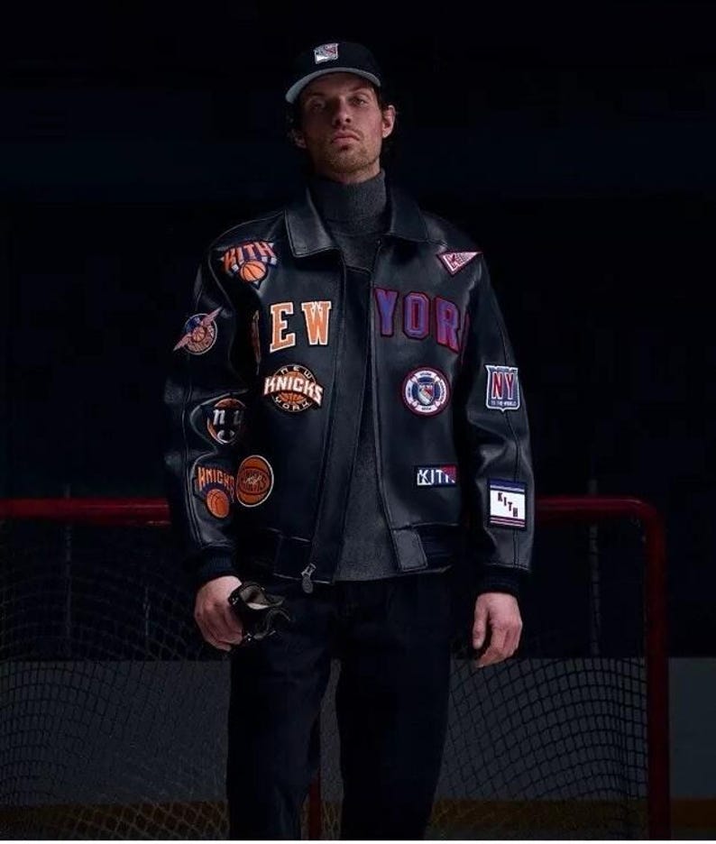 Op de afbeelding: Een zwart leren varsity-jack met diverse geborduurde patches, waaronder de tekst NEW YORK, KITH en Knicks-logo's. Het jack heeft een kraag, lange mouwen met geribbelde boorden en een ritssluiting aan de voorzijde over een grijze coltrui.