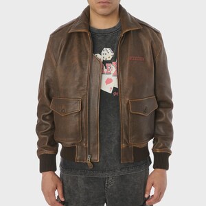 May include: Brown leather bomber jacket with a classic collar, two front pockets, and a ribbed hem and cuffs. The jacket features a zipper closure and a small patch on the chest with the word "AVIREX". The jacket is worn over a gray t-shirt.