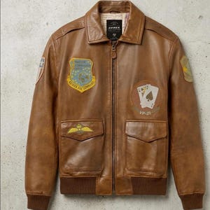 May include: Brown leather bomber jacket with multiple patches and pockets. The jacket features a zip-up front, a ribbed hem and cuffs, and a classic collar. Patches include the text "Strategic Reconnaissance Center" and a playing card design.