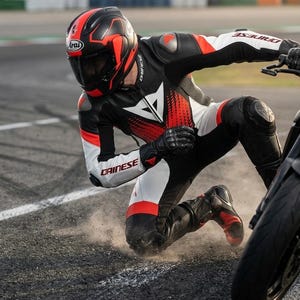 May include: A person wearing a black, white, and red motorcycle racing suit and helmet, kneeling near a motorcycle. The helmet has the brand name "Arai" on it. The suit has the brand name "Dainese" on it.
