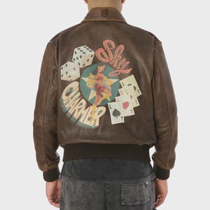 May include: Brown leather bomber jacket with a distressed finish. The back features a graphic with dice, playing cards, and a pin-up girl with the text "Sky Charmer". The jacket has a ribbed collar and cuffs.