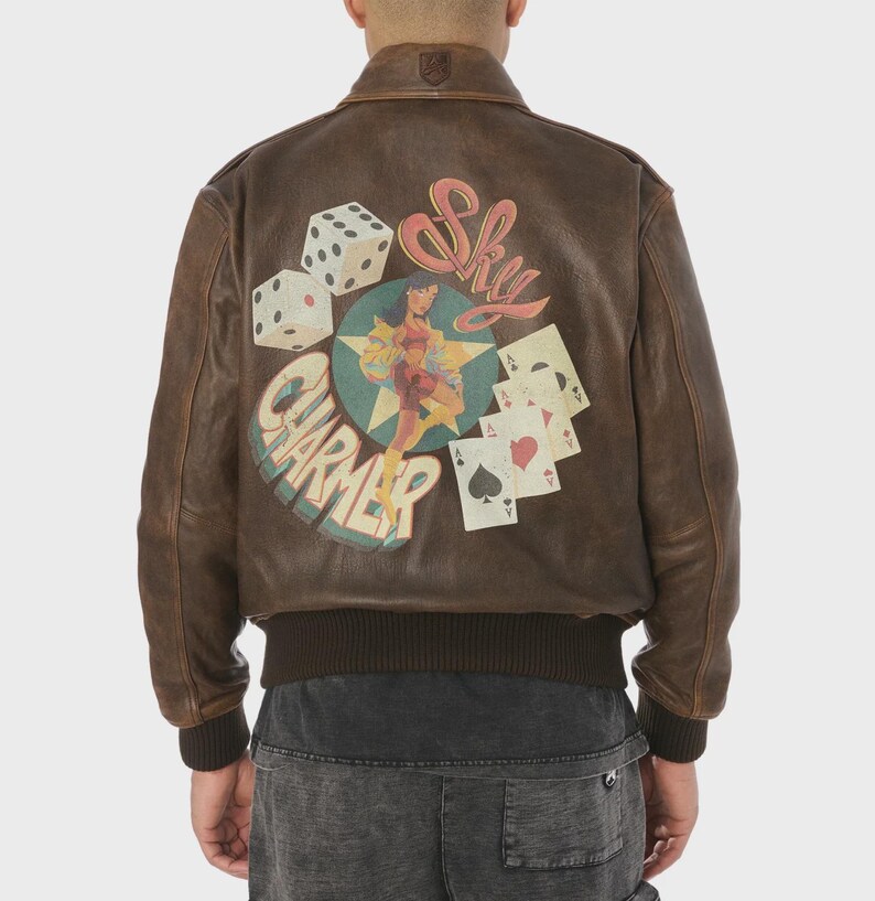 May include: Brown leather bomber jacket with a distressed finish. The back features a graphic with dice, playing cards, and a pin-up girl with the text "Sky Charmer". The jacket has a ribbed collar and cuffs.