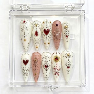 May include: A set of ten long, pointed artificial nails in a clear plastic case. The nails are primarily white and pink, with intricate designs including hearts, sunbursts, and ornate patterns. Embellishments include red and gold accents.