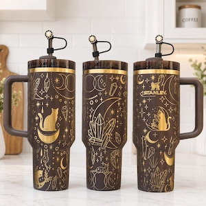 May include: Three dark brown Stanley tumblers with handles and lids, featuring gold-colored celestial and feline graphics including cats, crescent moons, stars, and crystals. Each tumbler has a black straw with a decorative topper attached.