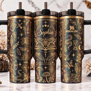 May include: Three black and gold stainless steel tumblers with handles and straws. The tumblers feature gold line art of moths, snakes, mushrooms, crystals, tarot cards, and hands. The center tumbler has the name Sophia printed in gold script.