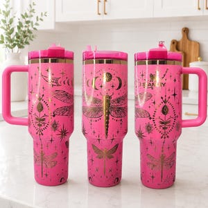 May include: Three bright pink Stanley tumblers with handles and lids, featuring gold-toned graphic prints of dragonflies, stars, and celestial moon phases. The tumblers have a metallic gold rim near the top and a consistent cylindrical shape.