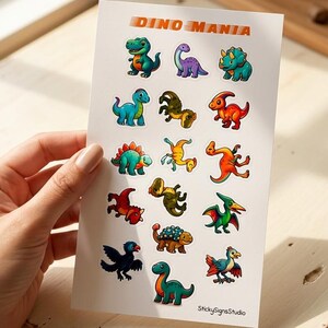 Könnte beinhalten: Ein weißer Aufkleberbogen mit dem Titel DINO MANIA, der fünfzehn bunte Cartoon-Dinosaurier-Illustrationen in verschiedenen Posen und Farben zeigt, darunter Grün, Blau, Orange und Violett. Unten rechts steht der Text StickySignsStudio.