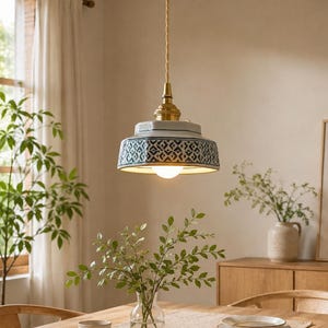 May include: A pendant light fixture featuring a ceramic shade with a blue and white geometric pattern, suspended by a gold-toned metal hardware and a fabric-covered cord, emitting a warm glow from a single light bulb centered underneath.