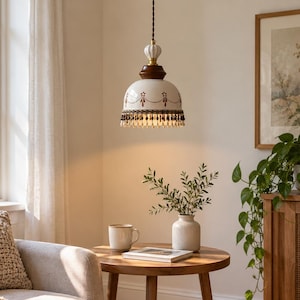 May include: A pendant light fitting with a white ceramic dome shade featuring a brown painted garland pattern and a beaded fringe trim. The lamp has a dark wood top section and a white fluted finial, suspended by a dark twisted fabric cord.