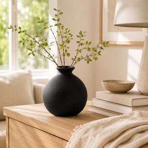 May include: A matte black spherical vase with a narrow neck, containing a branch with small green leaves. The vase is positioned on a light-colored wooden surface next to a stack of books, a small speckled bowl, and a lamp base.