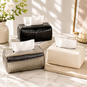 Puede incluir: Tres cajas rectangulares para pañuelos con una textura acanalada vertical. Una caja es negra brillante, otra es plateada metálica y la tercera es de color crema mate. Cada caja tiene una abertura central con pañuelos blancos sobresaliendo.