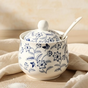 May include: A white ceramic sugar bowl with a lid and a small spoon. The bowl and lid are decorated with a blue floral pattern. The spoon is inserted into the bowl. The bowl is sitting on a white cloth.