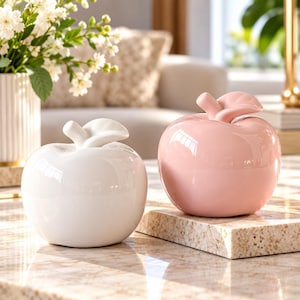 May include: Two smooth, glossy ceramic apple-shaped sculptures. One apple is solid white and the other is solid light pink. Each piece features a rounded body and a small, curved stem on top, reflecting light on their polished, reflective surfaces.