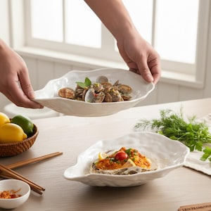 May include: Two white, leaf-shaped ceramic serving dishes, one held by hands, filled with seafood. The bottom dish contains a seafood dish with a tomato garnish. The top dish is filled with clams and herbs. The dishes are on a wooden table.